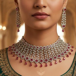 May include: A diamond necklace and matching earrings with ruby accents. The necklace is a choker style with a detailed design, and the earrings are long and dangling. The jewellery is set against a green embroidered garment.