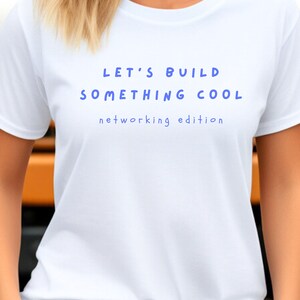 Könnte beinhalten: Ein weißes kurzärmeliges T-Shirt mit dem blauen Schriftzug LET'S BUILD SOMETHING COOL in einer fetten, abgerundeten Schriftart, gefolgt von dem kleineren blauen Text networking edition in einer einfachen, klaren serifenlosen Schrift.