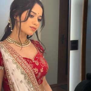 May include: A woman in a red and gold embroidered top and a white sari with gold trim. She is wearing a pearl necklace and earrings. The woman has dark hair and is looking down.