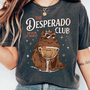 May include: Dark gray t-shirt with a graphic of a cat wearing sunglasses and a crown, holding a cocktail. The text reads "THE DESPERADO CLUB" and "SO FUN IT HURTS". Decorative stars are also included in the design.