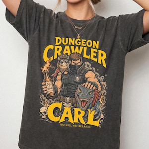 May include: A charcoal gray short-sleeve t-shirt featuring a graphic print with the text DUNGEON CRAWLER CARL and YOU WILL NOT BREAKME. The design shows a character holding a flaming bottle labeled JUG O' BOOM, a cat, and a dinosaur-like creature.