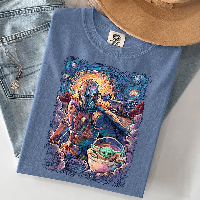 May include: A blue t-shirt featuring a colourful graphic print of a helmeted character and a small green creature in a floating pod. The artwork uses swirling, vibrant brushstrokes reminiscent of a starry night sky. The shirt has a Comfort Colors label.
