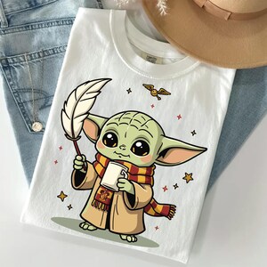 May include: A white t-shirt featuring a graphic of a green, large-eared creature wearing a tan robe and a red and yellow striped scarf. The creature holds a white quill and a white mug. Small stars and a winged golden ball are printed around the figure.