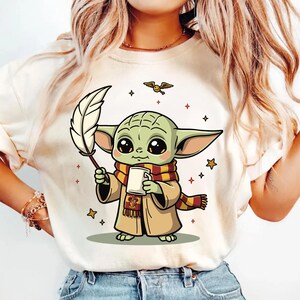 May include: A cream-colored t-shirt featuring a graphic of a small green creature with large eyes, wearing a tan robe and a red and yellow striped scarf. The creature holds a white quill and a mug, with a small golden snitch and stars nearby.