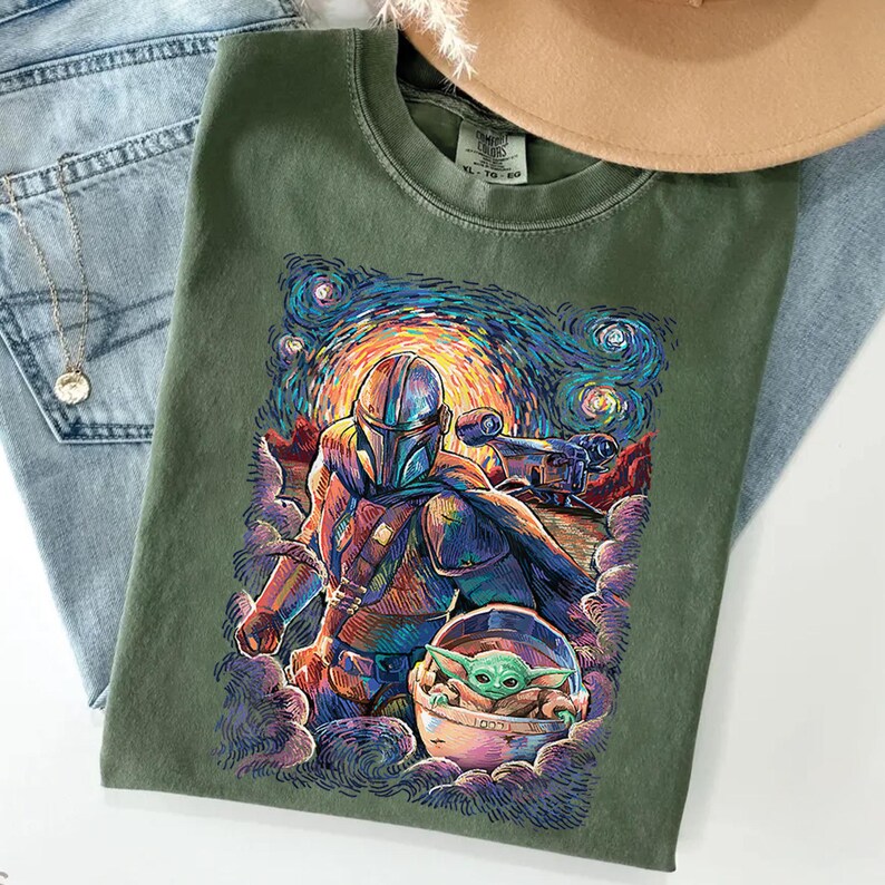 May include: An olive green crew neck t-shirt featuring a vibrant, swirling graphic print of a helmeted warrior and a small green creature in a floating pod. The artwork uses bold, colourful brushstrokes against a starry, night-sky background.