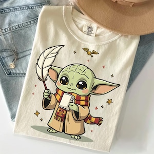 May include: A cream-colored t-shirt featuring a graphic of a green, large-eared creature wearing a red and yellow striped scarf and a tan robe. The character holds a white quill and a mug, with a small winged ball and stars surrounding it.