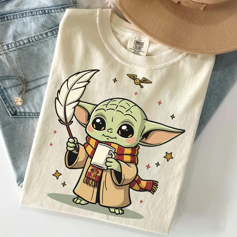 May include: A cream-colored t-shirt featuring a graphic of a green, large-eared creature wearing a red and yellow striped scarf and a tan robe. The character holds a white quill and a mug, with a small winged ball and stars surrounding it.
