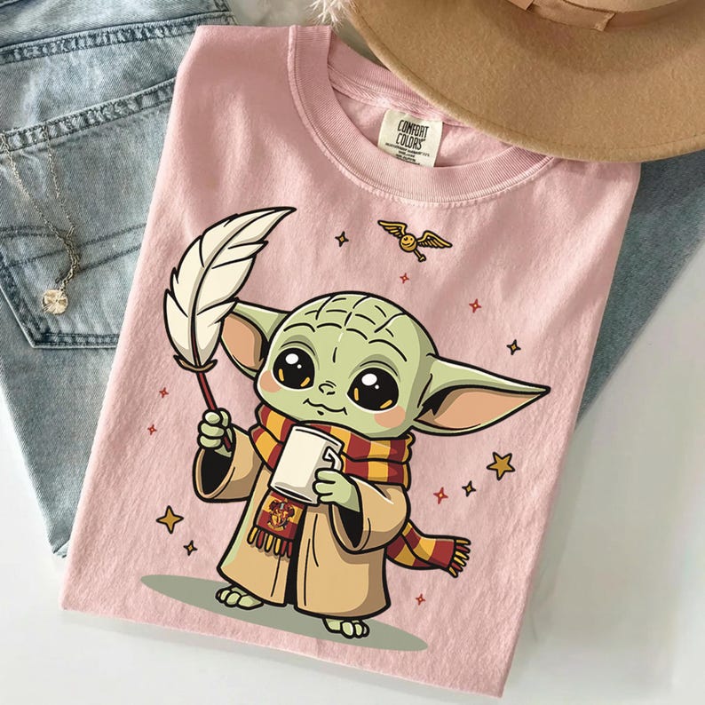 May include: A light pink t-shirt featuring a graphic of a green creature with large eyes, wearing a red and yellow striped scarf with a crest, holding a white mug and a white quill, surrounded by small stars and a flying winged golden ball.