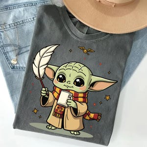 May include: A charcoal gray t-shirt featuring a cartoon illustration of a green creature with large eyes, wearing a tan robe and a red and yellow striped scarf. The character holds a white quill and a mug, with a small flying golden snitch above.