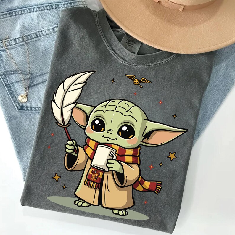 May include: A charcoal gray t-shirt featuring a cartoon illustration of a green creature with large eyes, wearing a tan robe and a red and yellow striped scarf. The character holds a white quill and a mug, with a small flying golden snitch above.