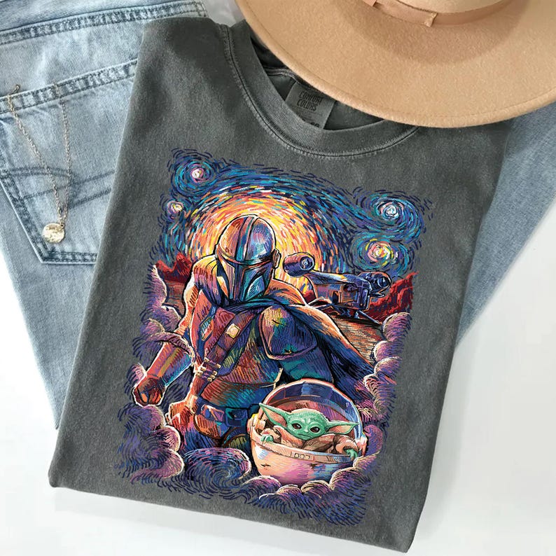 May include: A charcoal grey t-shirt featuring a colourful graphic print of a helmeted character in armour and a small green creature in a floating pod, set against a swirling, starry night sky background with vibrant blue, orange, and purple hues.