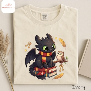 May include: Ivory-colored t-shirt featuring a cartoon dragon with a scarf, sitting on books, with an owl on a branch. The dragon has green eyes and black wings. Gold keys and stars are scattered around the design.