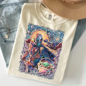 May include: A cream-coloured t-shirt featuring a colourful graphic print of a helmeted character in armour and a small green creature in a floating pod. The artwork uses swirling, textured brushstrokes in blue, orange, and purple hues.