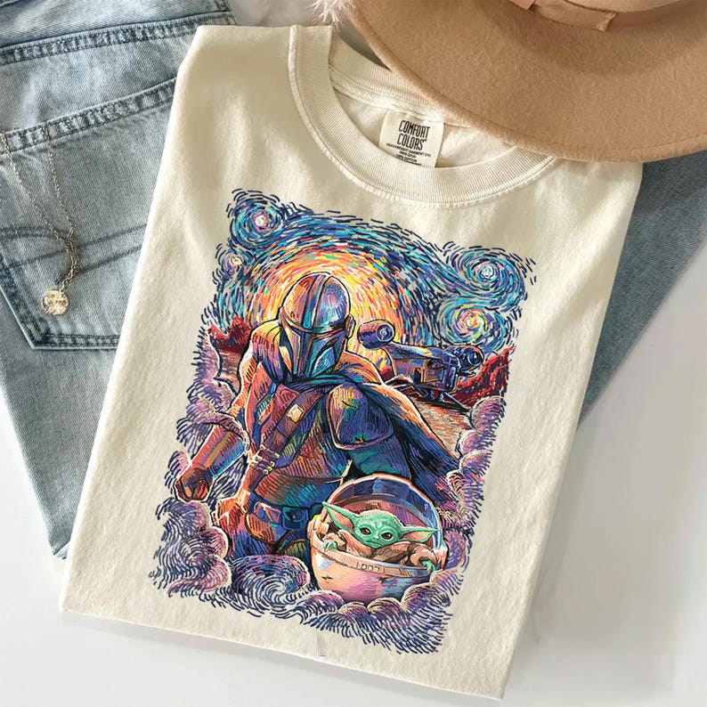May include: A cream-coloured t-shirt featuring a colourful graphic print of a helmeted character in armour and a small green creature in a floating pod. The artwork uses swirling, textured brushstrokes in blue, orange, and purple hues.