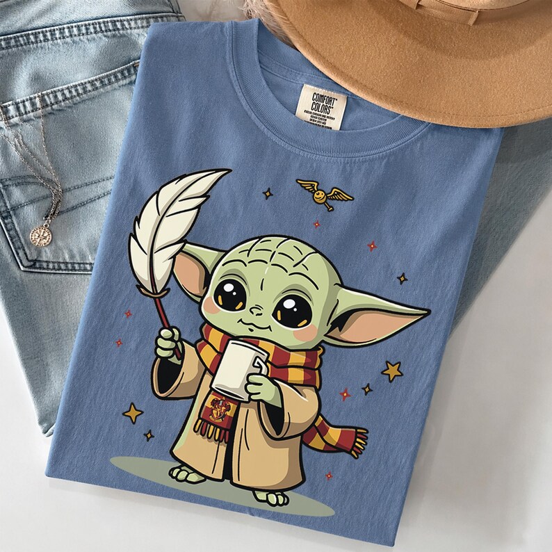 May include: A blue t-shirt featuring a cartoon illustration of a green creature with large eyes, wearing a tan robe and a red and yellow striped scarf. The creature holds a white quill and a mug. Small stars and a winged golden ball surround it.