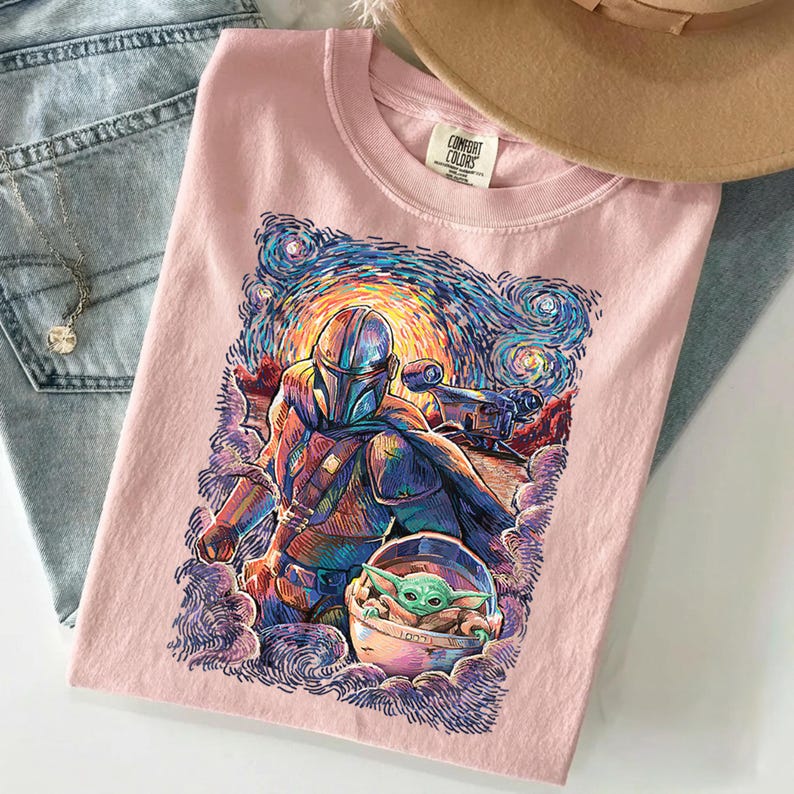 May include: A light pink t-shirt featuring a graphic print of an armoured character and a small green creature in a floating pod. The artwork uses swirling, colourful brushstrokes reminiscent of a starry night sky. The shirt has a Comfort Colors label.