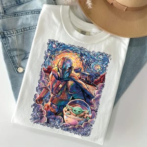May include: A white t-shirt featuring a colourful graphic print of a helmeted character in armour and a small green creature in a floating pod. The artwork uses swirling brushstroke patterns in shades of blue, orange, and purple, reminiscent of a night sky.