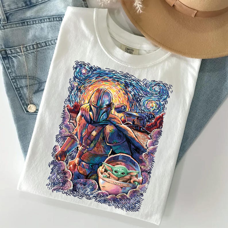 May include: A white t-shirt featuring a colourful graphic print of a helmeted character in armour and a small green creature in a floating pod. The artwork uses swirling brushstroke patterns in shades of blue, orange, and purple, reminiscent of a night sky.