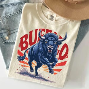May include: A cream-colored t-shirt featuring a graphic of a dark blue buffalo running in front of a red sunburst design. The word BUFFALO is printed in large red letters arching over the animal. The shirt has a Comfort Colors brand label.