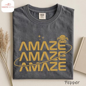 May include: A dark gray t-shirt with the word "AMAZE" stacked three times in a gold color, with a graphic of a planet and stars. The shirt has a crew neck and a tag that says "Comfort Colors". The word "Pepper" is at the bottom right.
