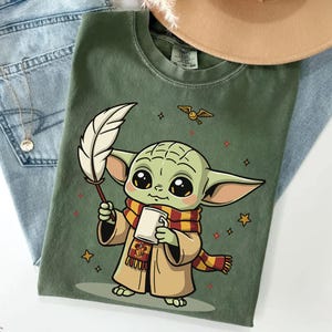 May include: An olive green t-shirt featuring a graphic of a small green creature with large eyes, wearing a tan robe and a red and yellow striped scarf. The character holds a white quill and a mug, with a small winged golden ball and stars nearby.
