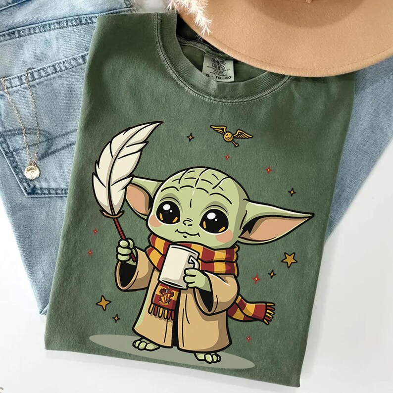 May include: An olive green t-shirt featuring a graphic of a small green creature with large eyes, wearing a tan robe and a red and yellow striped scarf. The character holds a white quill and a mug, with a small winged golden ball and stars nearby.