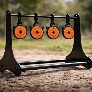 Op de afbeelding: Een zwarte stalen doelstandaard met vier oranje en zwarte doelen. Elk doel heeft een bullseye-ontwerp. De standaard staat buiten op een vuil oppervlak met een vage achtergrond van bomen en gras.