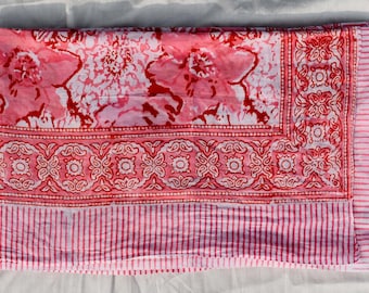 May include: A folded pink and white floral patterned textile. The design features large pink flowers and a border with a repeating geometric pattern. The bottom edge has pink and white stripes.