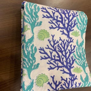 Pode incluir: Uma peça de tecido dobrada com fundo branco e um padrão repetitivo de coral azul-escuro, algas verde-azuladas e conchas em espiral verde-claras. A estampa está distribuída pela superfície em um arranjo denso com tema náutico.
