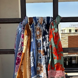 Könnte beinhalten: Eine Sammlung von fünf rechteckigen Schals, die über einen dunklen Metallrahmen drapiert sind. Die Schals weisen verschiedene Muster auf, darunter Blumen-, Mandala- und geometrische Designs in einer Farbpalette aus Blau, Orange, Braun, Weiß und Salbeigrün.