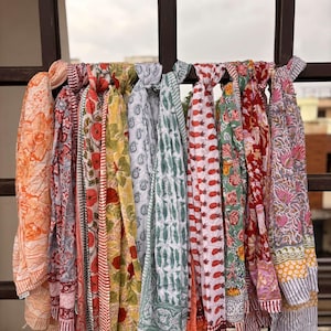 May include: A display of patterned scarves in a range of colours, including orange, red, green, yellow, and pink, hanging on a dark brown metal rack. The scarves showcase floral and geometric designs. The fabric appears lightweight and airy, suitable for warmer weather.