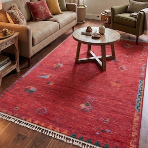 Puede incluir: Un salón con una alfombra roja con diseños geométricos, un sofá marrón con cojines y una mesa de centro de madera. Un sillón verde y una planta en maceta añaden color. Una lámpara y grabados botánicos enmarcados completan el acogedor interior.