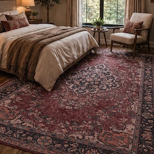 May include: A large rectangular area rug featuring a distressed floral pattern in shades of deep red, charcoal gray, and muted pink. The rug has a wide decorative border with intricate botanical motifs and sits on a wooden floor in a bedroom.
