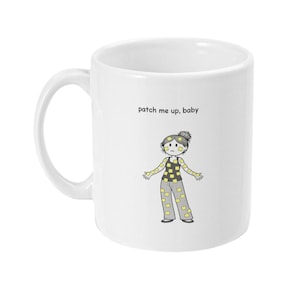 Może przedstawiać: Biały ceramiczny kubek z kreskówkową ilustracją osoby w patchworkowych ubraniach. Tekst "patch me up, baby" jest wydrukowany nad ilustracją. Kubek ma zakrzywiony uchwyt.