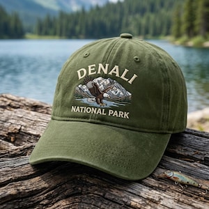 Könnte beinhalten: Eine olivgrüne Baseballkappe mit einer Stickerei, die einen Weißkopfseeadler zeigt, der über ein Gebirge und einen See fliegt. Der Schriftzug "DENALI" ist bogenförmig darüber angeordnet, während "NATIONAL PARK" in einer Zeile darunter steht.