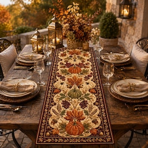 Puede incluir: Un camino de mesa rectangular con un estampado otoñal detallado de calabazas naranjas, bellotas marrones, espigas de trigo y hojas coloridas en tonos rojos, dorados y verdes, con un borde rojo oscuro sobre una mesa de madera rústica.