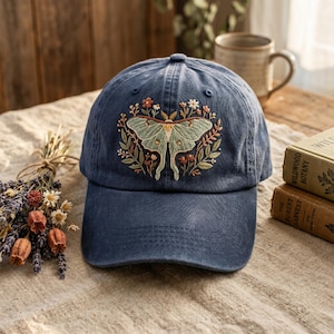 May include: A navy blue baseball cap with an embroidered design of a green moth surrounded by flowers and leaves. The cap is made of a soft fabric and has a curved brim. The design is detailed and colorful, with shades of green, brown, and orange.