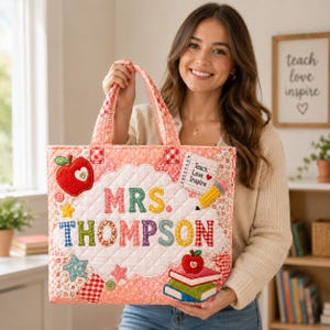 Puede incluir: Un bolso tote acolchado en tonos rosa y estampados florales con el texto MRS. THOMPSON en letras de tela coloridas. Los apliques incluyen una manzana roja, un bloc de notas con el texto Teach Love Inspire, un lápiz y libros.