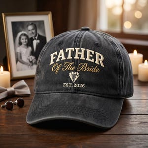 Puede incluir: Gorra de béisbol negra con el texto "FATHER Of The Bride" en letras blancas y doradas. La gorra también presenta un gráfico de esmoquin y el año "EST. 2026". Una foto en blanco y negro enmarcada está en el fondo.