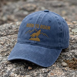 Puede incluir: Una gorra de béisbol azul con acabado desgastado que presenta un diseño bordado en hilo dorado. El bordado muestra un lobo aullando sobre una roca frente a picos montañosos, con el texto BORN TO ROAM situado sobre el lobo.
