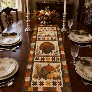 May include: A rectangular fabric table runner featuring a patchwork border in shades of orange, brown, and green. The centre panel displays printed illustrations of white pumpkins, a central turkey, and a harvest arrangement of corn and gourds.