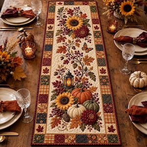 Può includere: Un runner da tavola trapuntato rettangolare con un pannello centrale color crema stampato con girasoli, zucche, una lanterna, bacche, foglie autunnali e una farfalla, bordato da un motivo patchwork di quadrati a quadri e floreali.