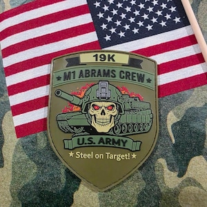 May include: An olive green patch featuring a stylized M1 Abrams tank with a skull-faced soldier in a helmet. The patch reads "19K M1 ABRAMS CREW U.S. ARMY Steel on Target!" An American flag is in the background.