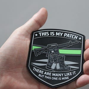 May include: A shield-shaped black tactical patch featuring a rifle, a pair of boots, and dog tags. The text reads THIS IS MY PATCH and THERE ARE MANY LIKE IT BUT THIS ONE IS MINE. A thin horizontal neon green line runs across the center of the patch.