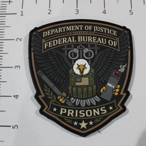 May include: A shield-shaped patch featuring a bald eagle wearing a tactical vest with an American flag, holding a baton and an MK4 pepper spray canister. The text reads DEPARTMENT OF JUSTICE FEDERAL BUREAU OF PRISONS. A ruler shows it is 3 inches tall.