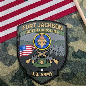 May include: A military patch with the text "FORT JACKSON SOUTH CAROLINA" and "U.S. ARMY". The patch features a torch, crossed rifles, and silhouettes of soldiers. An American flag is in the background.