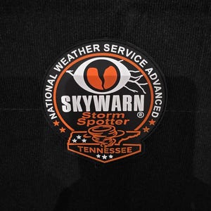 May include: A circular patch with the text "NATIONAL WEATHER SERVICE ADVANCED SKYWARN Storm Spotter TENNESSEE" in white and orange. The patch features a stylized eye and a tornado graphic on a black background.