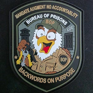 May include: A shield-shaped morale patch featuring a cartoon eagle wearing a cap and uniform, holding a set of keys. The text reads MANDATE AUGMENT NO ACCOUNTABLITY, BUREAU OF PRISONS, BOP, and BACKWORDS ON PURPOSE. The design includes a prison tower.