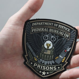 May include: A shield-shaped patch featuring the text DEPARTMENT OF JUSTICE FEDERAL BUREAU OF PRISONS. The design includes a bald eagle wearing a tactical vest with a flag patch, holding a baton and an MK4 pepper spray canister, with handcuffs above.