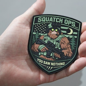 May include: A shield-shaped morale patch featuring a brown Bigfoot character in tactical gear with night vision goggles and a rifle. The background shows a forest, a crescent moon, and a stylized American flag. Text reads SQUATCH OPS and YOU SAW NOTHING.